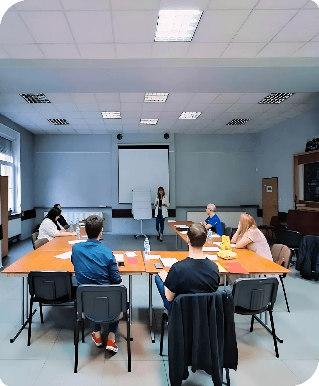 Grupa osób na sali konferencyjnej, kobieta prowadzi prezentację przy tablicy.