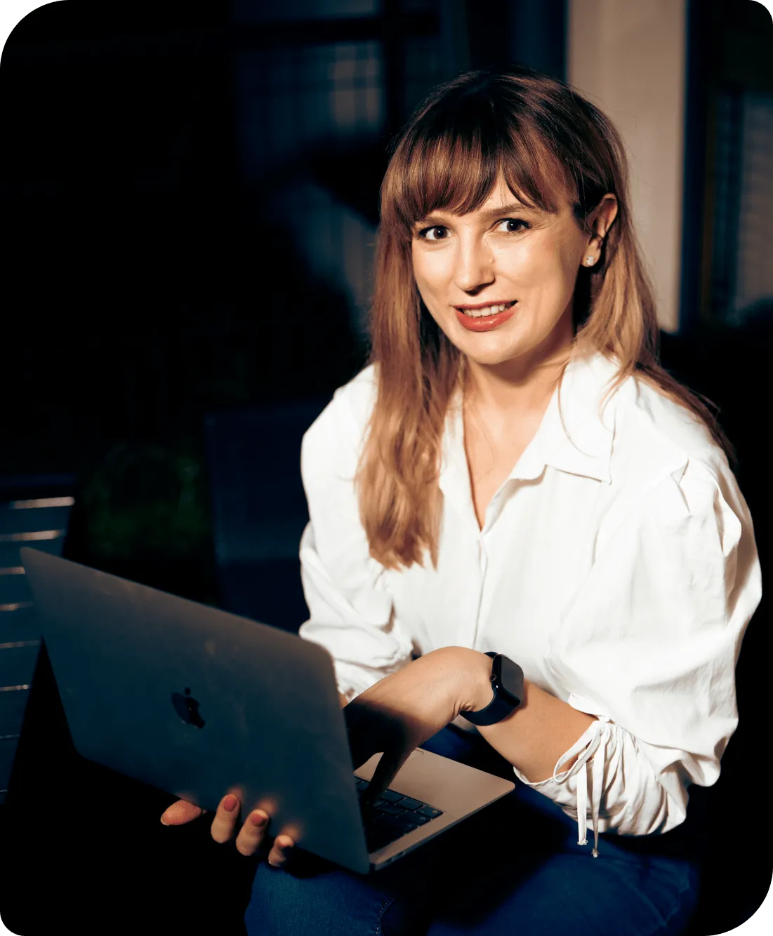 Kobieta siedząca przy stole, trzymająca laptopa i uśmiechająca się, w białej koszuli.